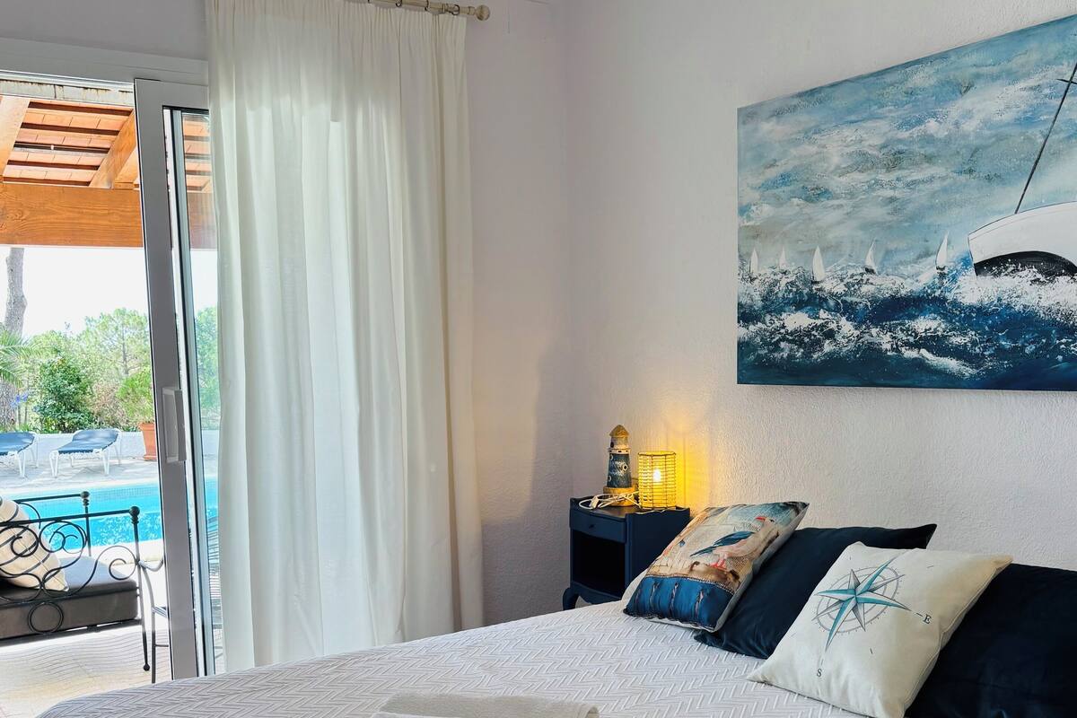 Une chambre d’hôtel avec un lit, un tableau représentant un bateau sur l’eau, une table de chevet avec une lampe et une vue sur une piscine à l’extérieur.