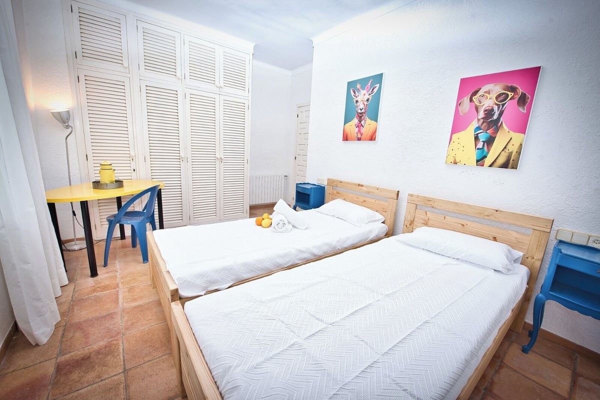 Une chambre avec deux lits, une petite table, une chaise et des œuvres d’art accrochées aux murs.