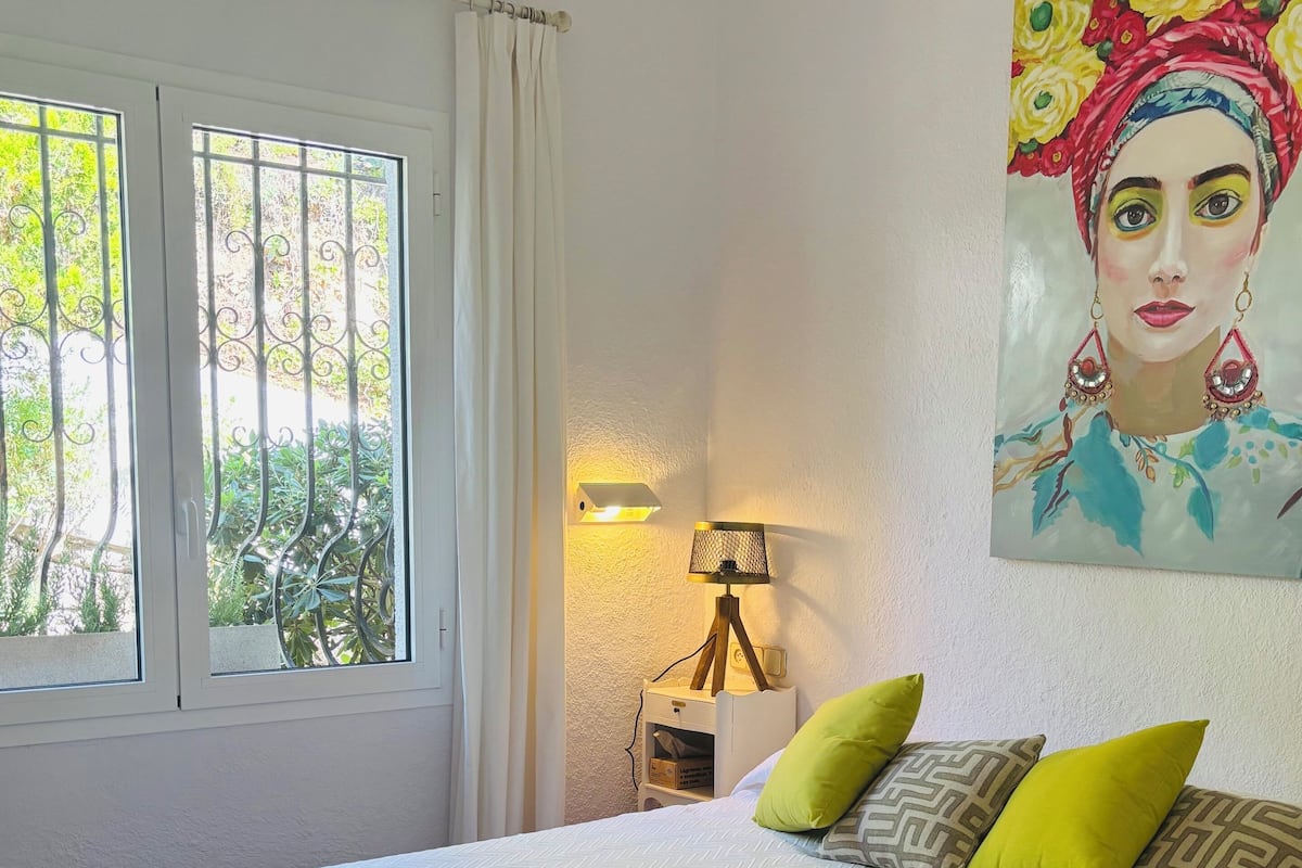 Un lit bien fait, recouvert d’une courtepointe blanche, avec des oreillers verts à motifs et une serviette blanche pliée posée sur le lit. Un tableau est accroché au mur au-dessus du lit, ainsi qu’une lampe de chevet et une fenêtre avec des rideaux.