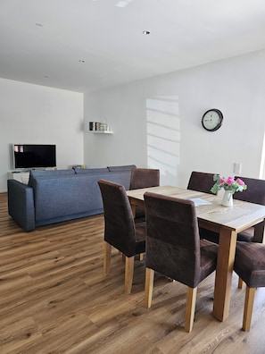 Ein modernes Wohnzimmer mit Sofa, Esstisch, Stühlen, einem Flachbildfernseher, einer Uhr und einem Bücherregal.