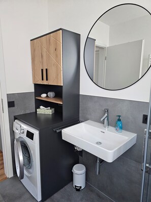Ein kleines Badezimmer mit Waschmaschine, Waschbecken, Spiegel und einem kleinen Mülleimer.