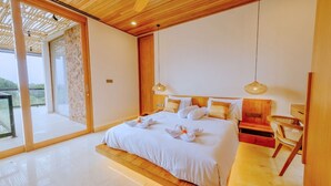 A modern bedroom with a large bed, wooden headboard, two bedside lamps, and a view through a glass door to an outdoor area. - D'Kamboja KIS1 Managed by D'Kamboja Group (Bali)