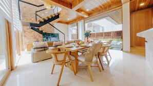 A spacious living area with a wooden dining table, wicker chairs, and a sofa. A staircase with a glass railing leads to an upper level. - D'Kamboja KIS1 Managed by D'Kamboja Group (Bali)