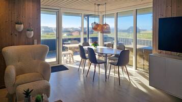 Un salon moderne doté d’une grande fenêtre, d’un coin repas avec une table et des chaises, et d’un téléviseur posé sur un meuble.