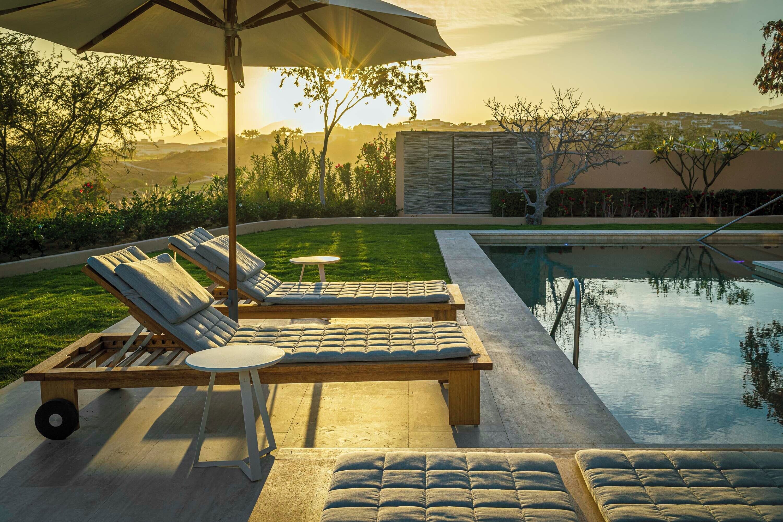 Un espace aménagé au bord de la piscine, avec des chaises longues et une table placée sous un parasol, le tout entouré d’une pelouse bien entretenue et offrant une vue sur les collines.