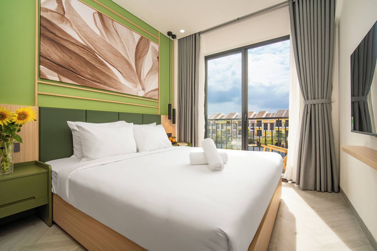 Habitación de hotel con una cama grande, pared decorativa verde, un ventanal con cortinas y vistas a edificios.