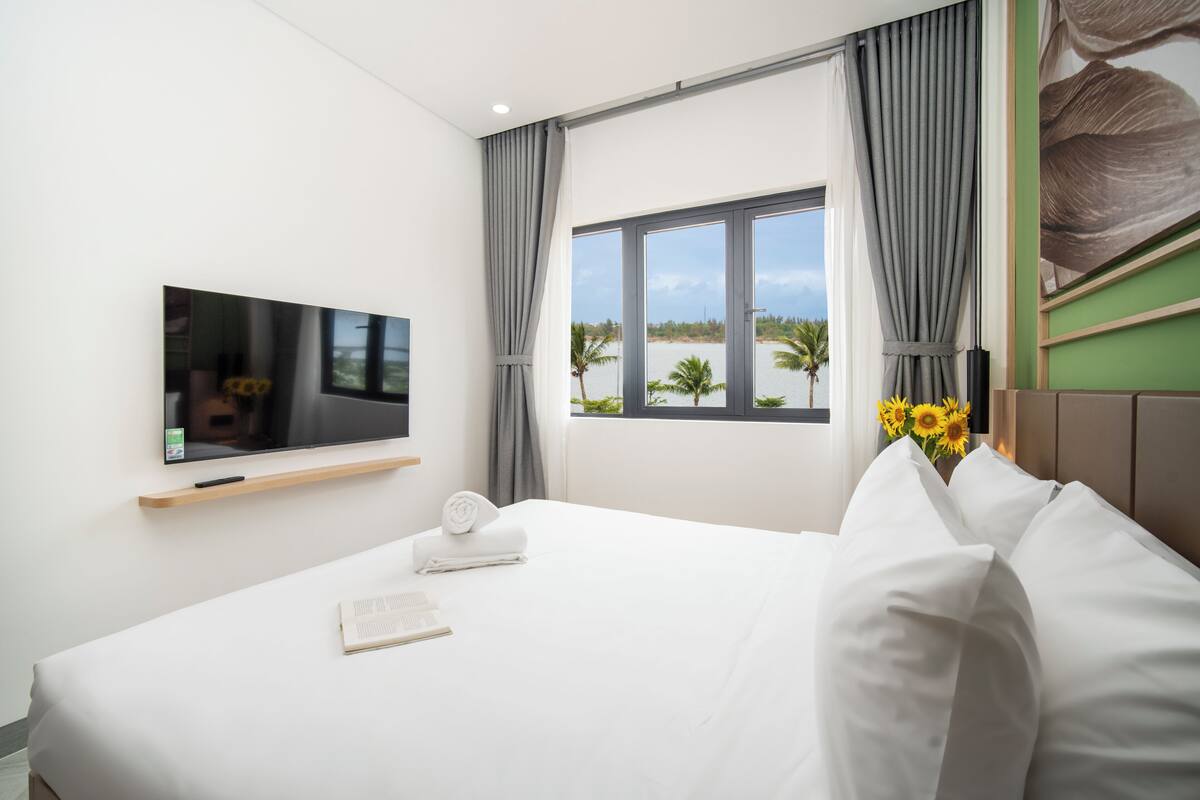 Habitación de hotel con cama, televisor en la pared, ventana con vista a palmeras y un florero con flores.