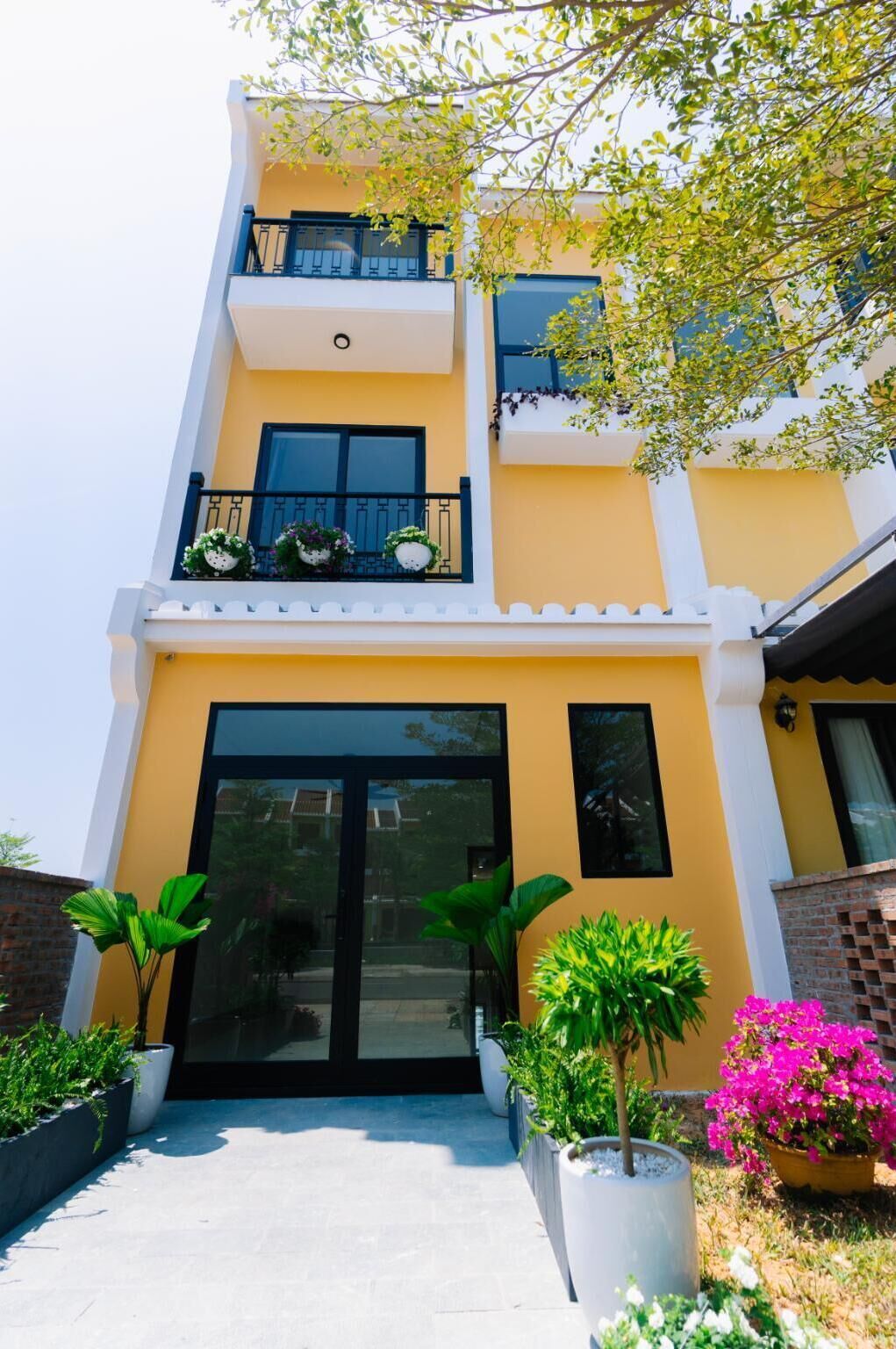Un immeuble de deux étages avec une façade jaune, un balcon orné de plantes en pot et une grande entrée vitrée.