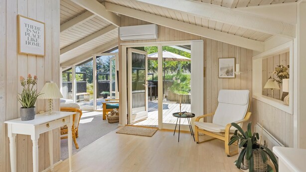 A spacious room with a high ceiling, a large sliding glass door leading to an outdoor deck, a white console table with drawers, a wicker chair, and a small round table. - 3 bedroom beautiful home in Blokhus (Blokhus)