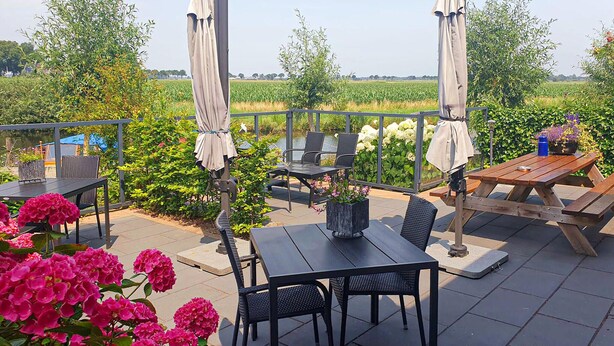 Een terras buiten met tafels en stoelen, omringd door groen en uitzicht op velden.