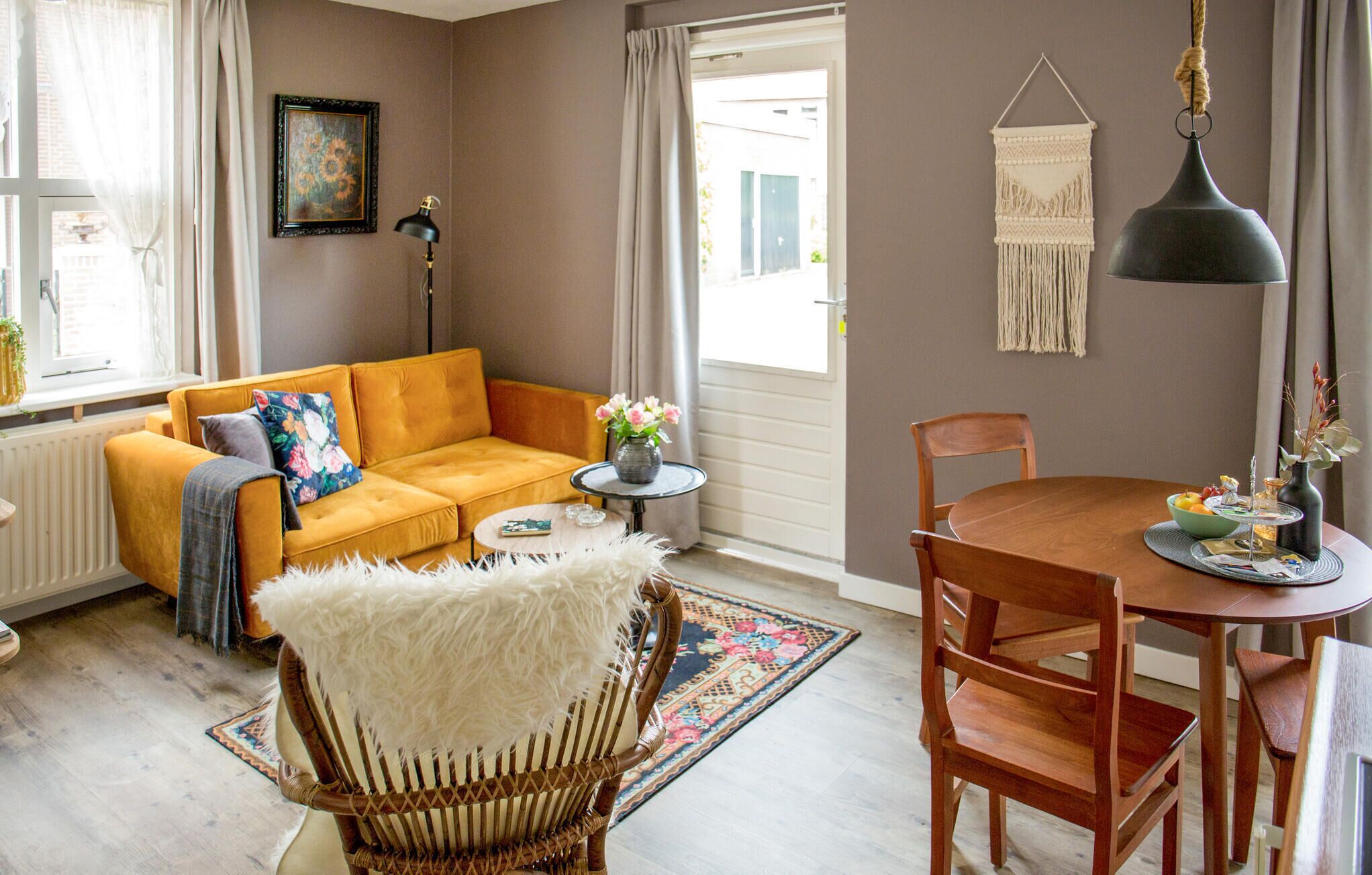 Ein gemütliches Wohnzimmer mit einem senfgelben Sofa, einem weißen, flauschigen Sessel, einem Holzespeisetisch mit Stühlen, einem kleinen Beistelltisch mit Blumen in einer Vase und einer Wandbehagung aus Makramee.