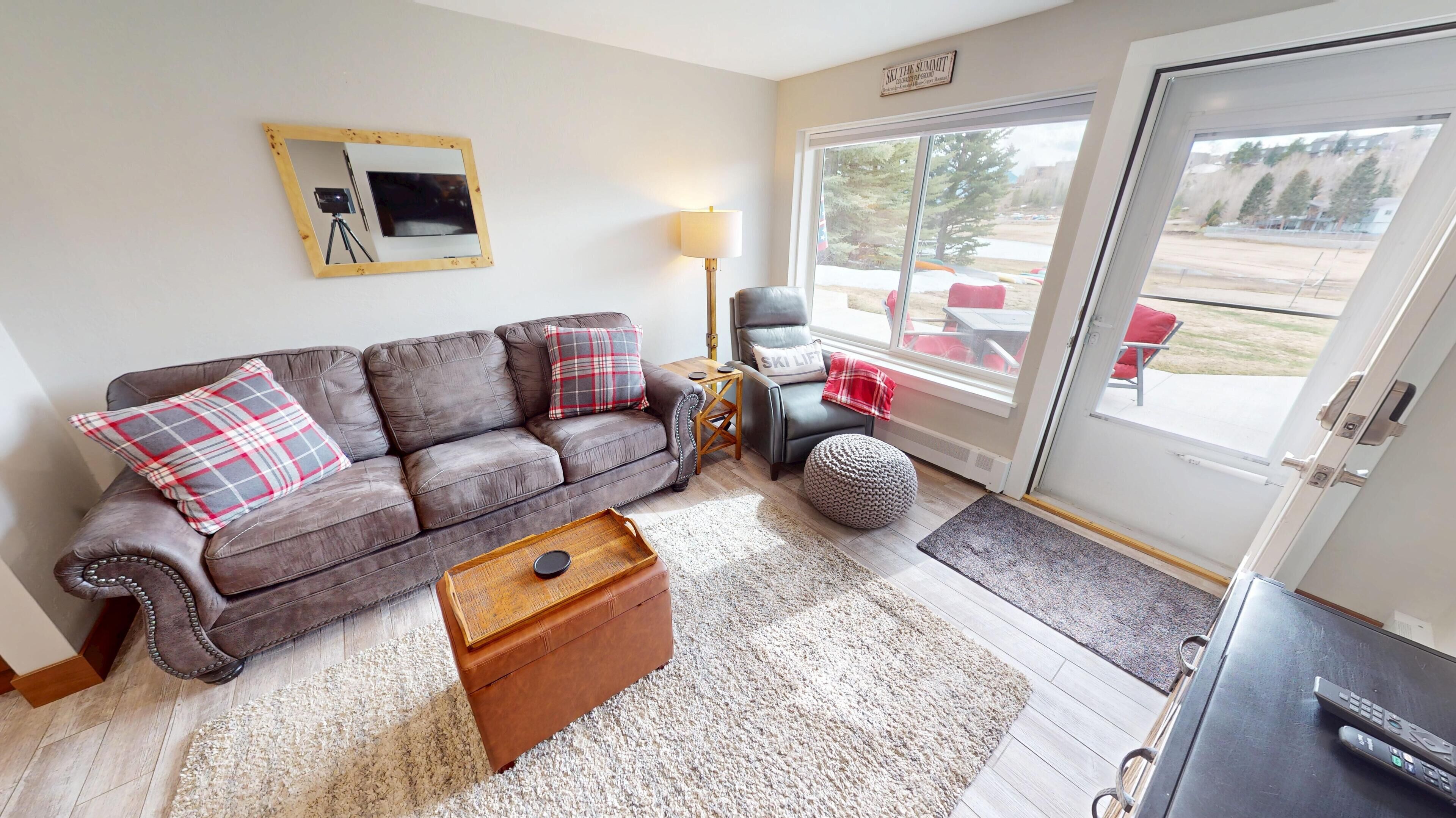 A cozy living room with a brown leather sofa, a wooden coffee table, and a large window offering a view of outdoor seating.