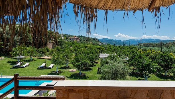 A poolside view with a thatched roof, lounge chairs, and a view of a lush landscape with trees and distant mountains. - Neriana Villa - Garden Paradise (Chania)