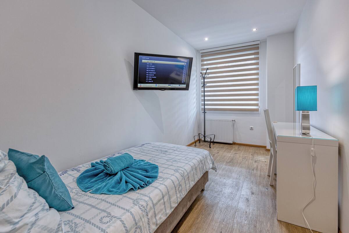 Ein Schlafzimmer mit Bett, Wandfernseher, Fenster mit Jalousien und Schreibtisch mit Lampe.