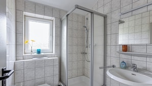 Ein Badezimmer mit einer gläsernen Duschkabine, einem weißen Waschbecken und einem Fenster mit Blumen in einer Vase.