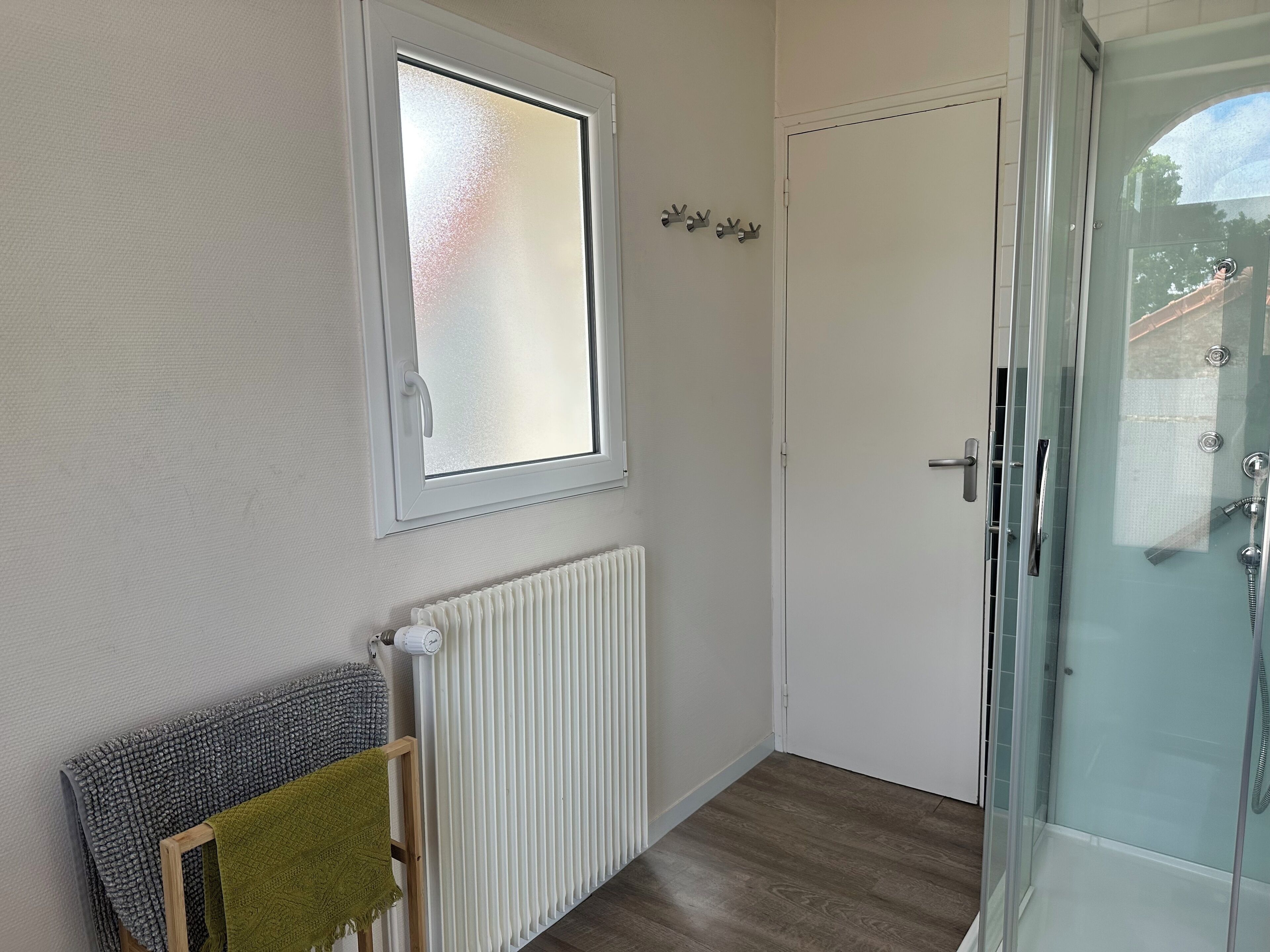 Un baño con mampara de vidrio, radiador blanco, ventana con cristal satinado y un perchero con una toalla verde.