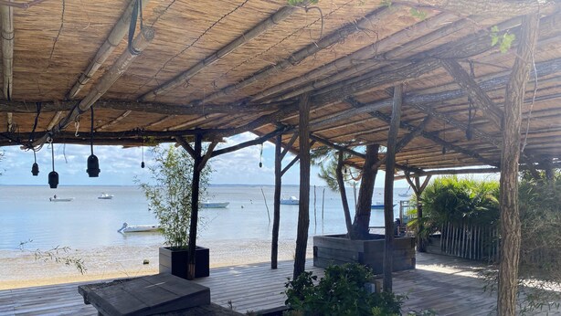 Una terraza de madera con techo de paja, con vista a un mar tranquilo y barcos.