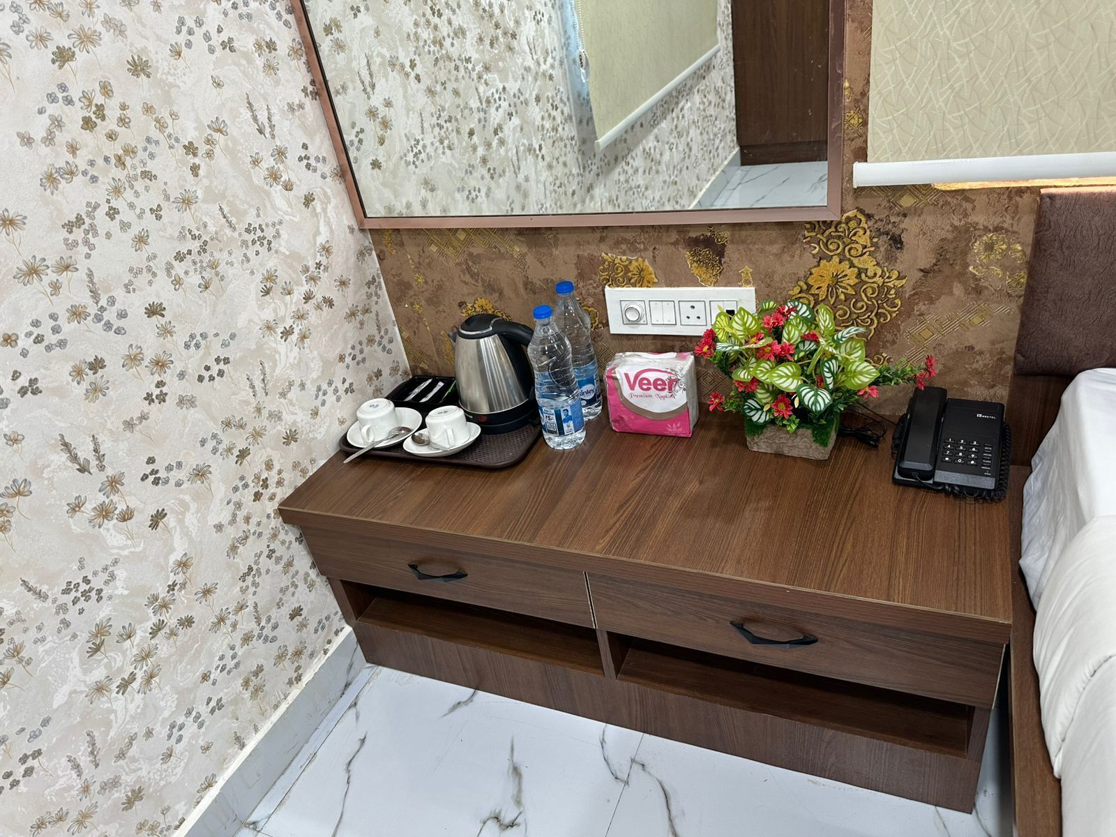 Une chambre d’hôtel avec une table de chevet en bois, un miroir, un papier peint à motifs floraux, une petite plante, une bouteille d’eau, une boîte de mouchoirs, une bouilloire, un grille-pain et un téléphone.