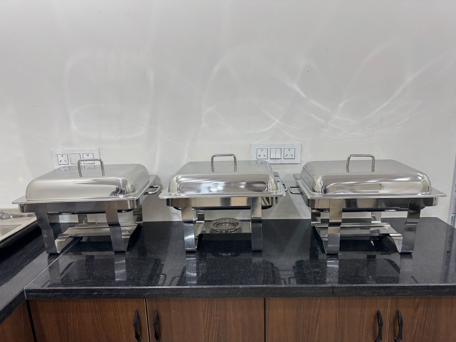 Three stainless steel buffet covers on a black countertop with wooden cabinets below.