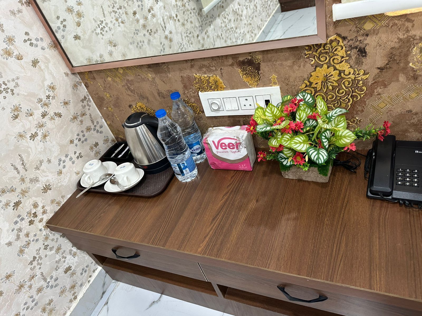 Et træbord med elkedel, vandflasker, tissues, en telefon og en pyntplante.
