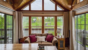 A cozy room with a wooden ceiling, large windows, a wooden table, and a cushioned seating area.