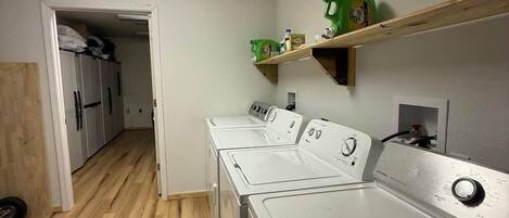 Un lavadero con piso de madera, lavadoras blancas y un estante con productos de limpieza.