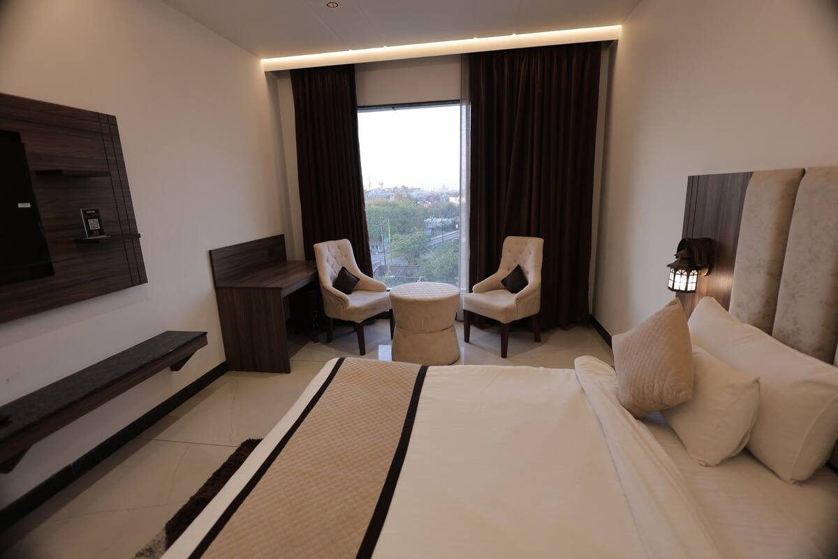 A hotel room with a large bed, two armchairs, a small table, a TV mounted on the wall, and a window with curtains.