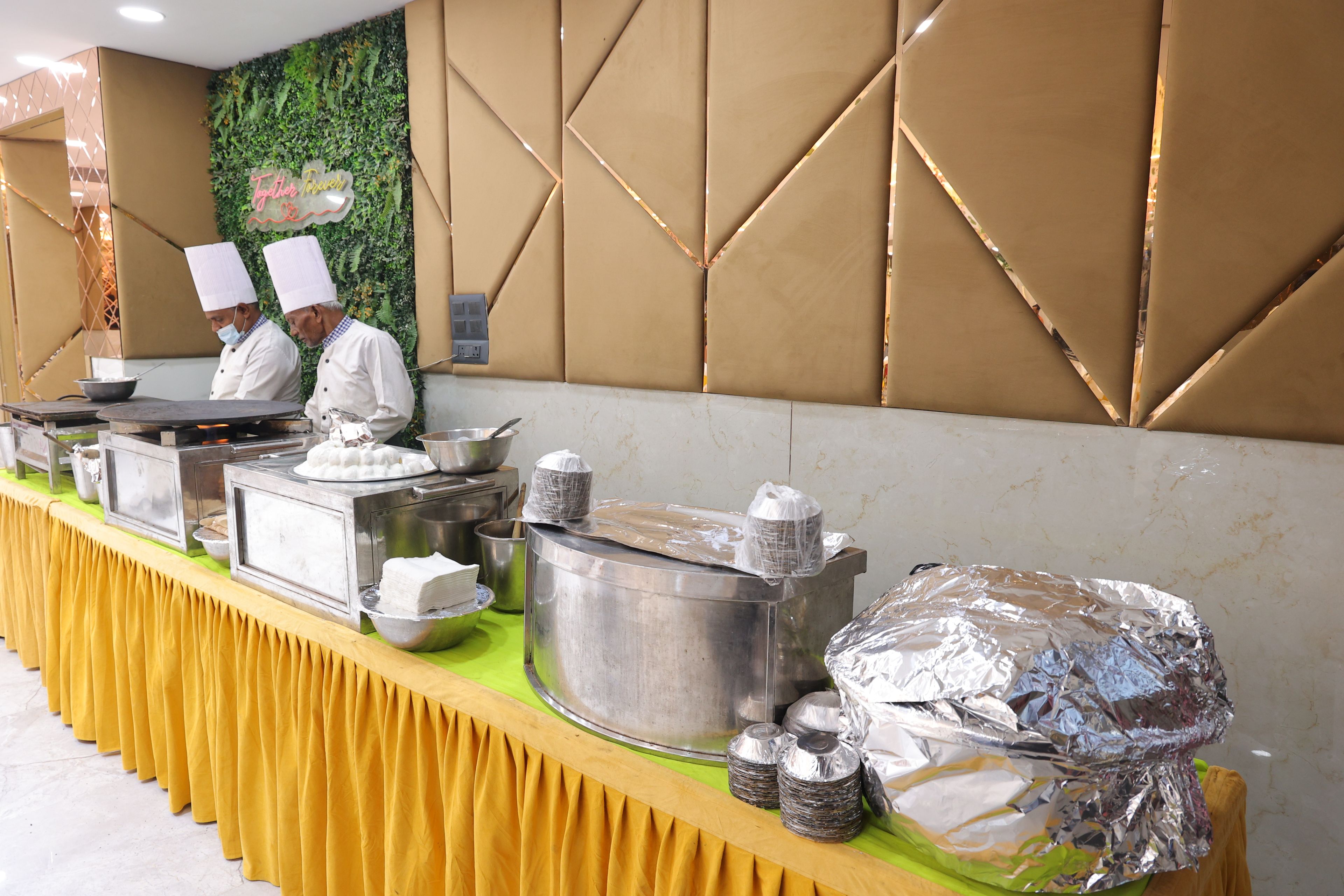 A buffet setup with food service staff, a green wall with a floral design, and a patterned wall in the background.