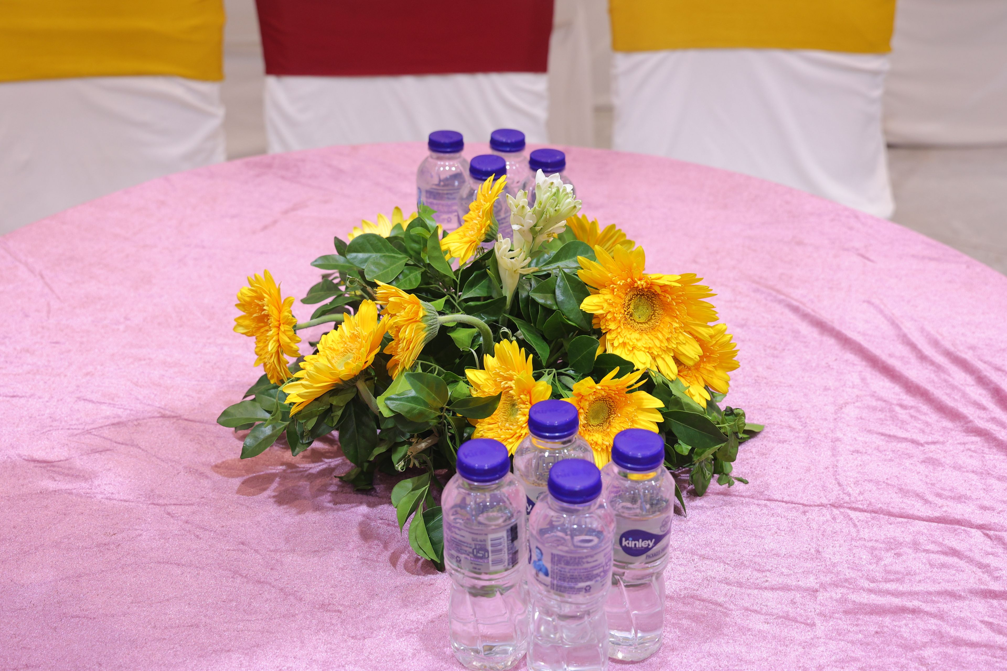 A round table with a pink tablecloth, surrounded by yellow flowers and clear plastic bottles with purple caps.