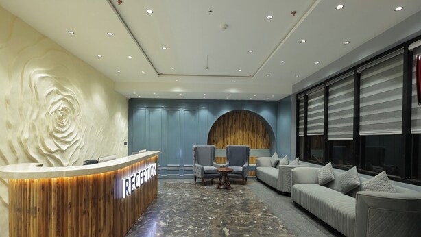 A reception area with a wooden reception desk, a grey sofa, and a seating area with armchairs. - SNJ TAJ GRAND HOTEL (Agra)