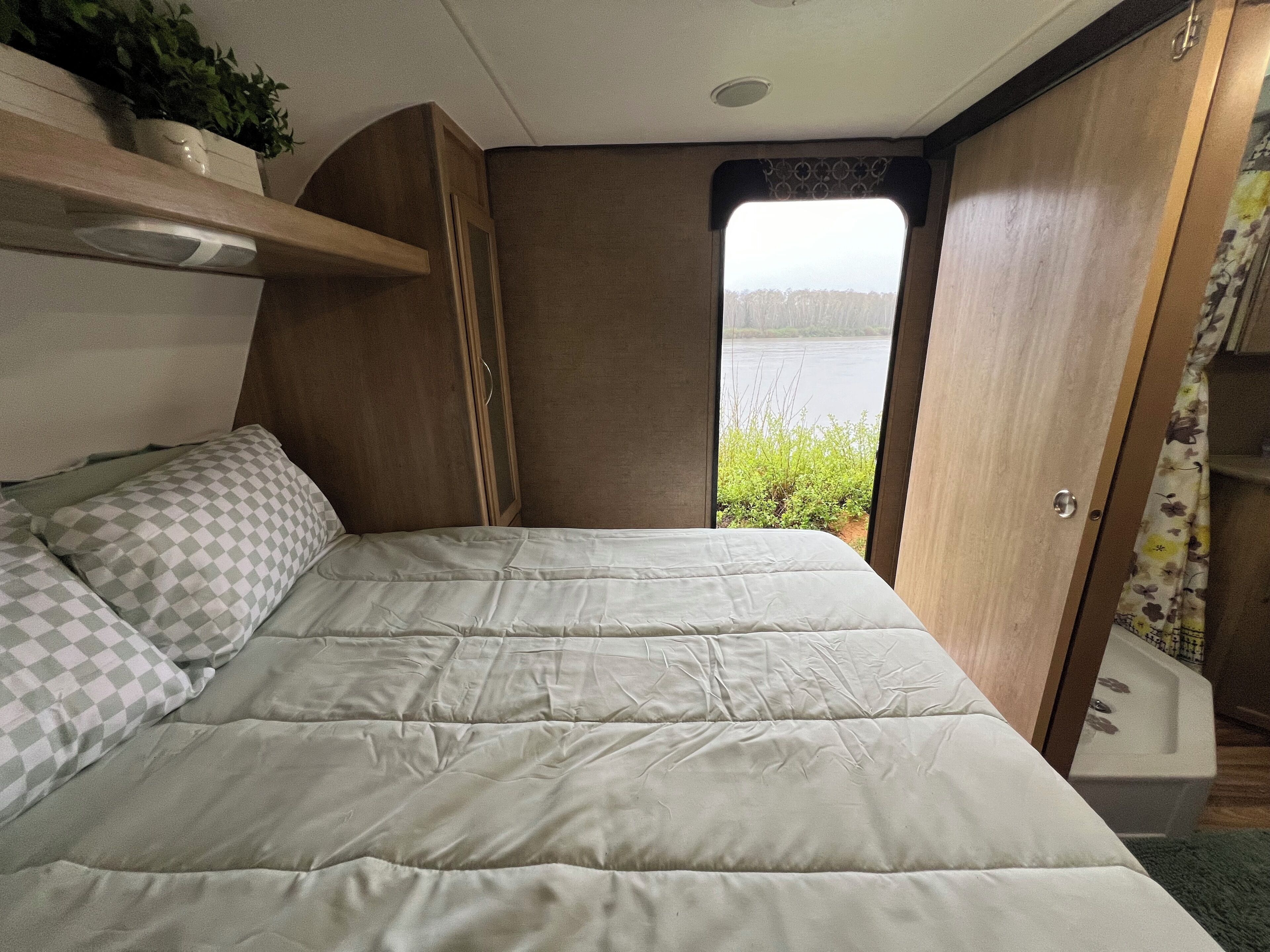 A bed with a checkered pillow, a wooden wardrobe, and a view of a lake through a window.