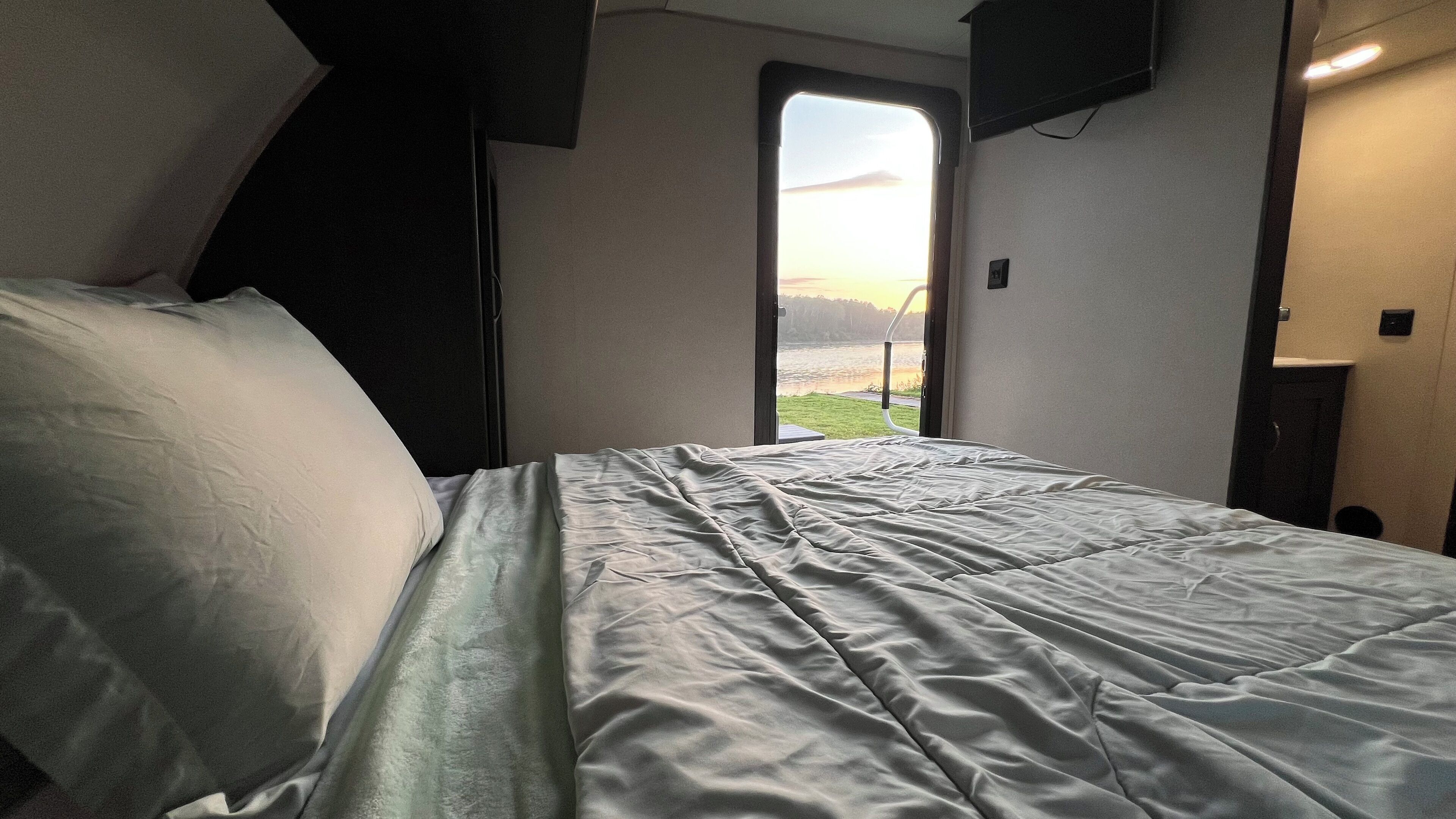 A bed with a view of a body of water and a sunset through a window.