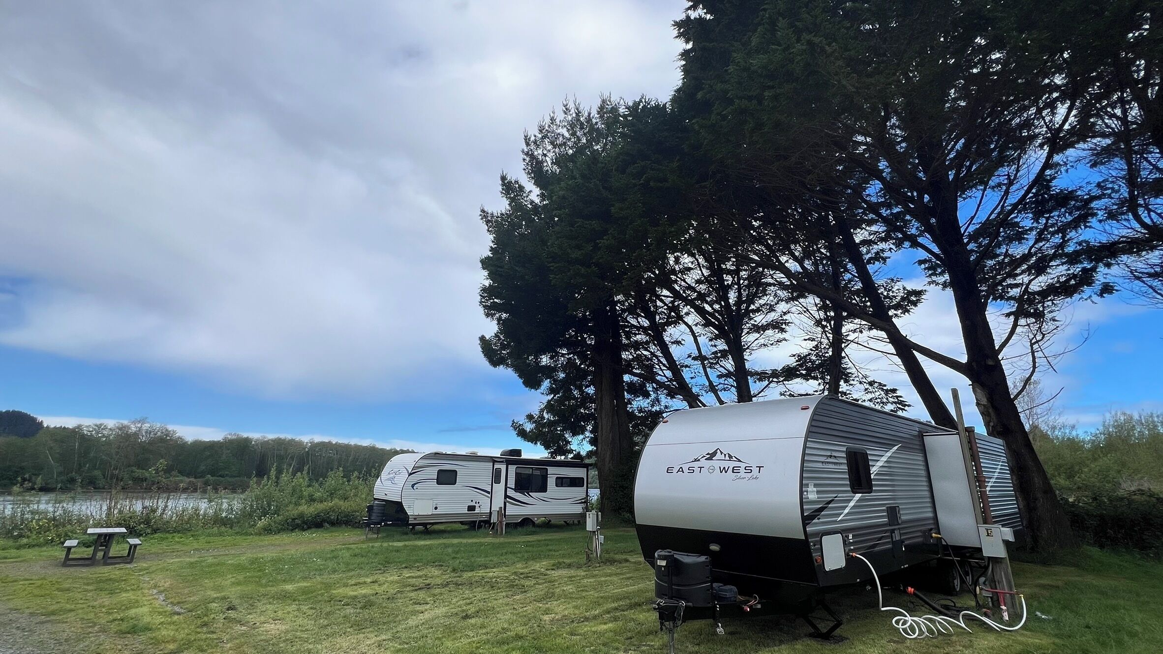 Two RVs parked in a grassy area with trees and a lake in the background.
