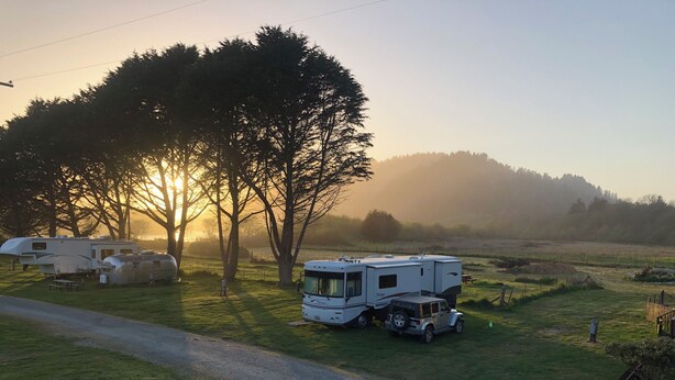 A campsite with a caravan and a small SUV parked near a dirt road, surrounded by trees and open fields.