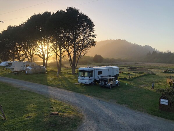 A campsite with a caravan and a small SUV parked near a dirt road, surrounded by trees and open fields.