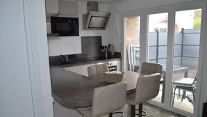 A modern kitchen with a built-in microwave, oven, and sink. There is a dining area with a table and chairs, and a sliding glass door leading to an outdoor patio. - Country cottage (Raon-aux-Bois)