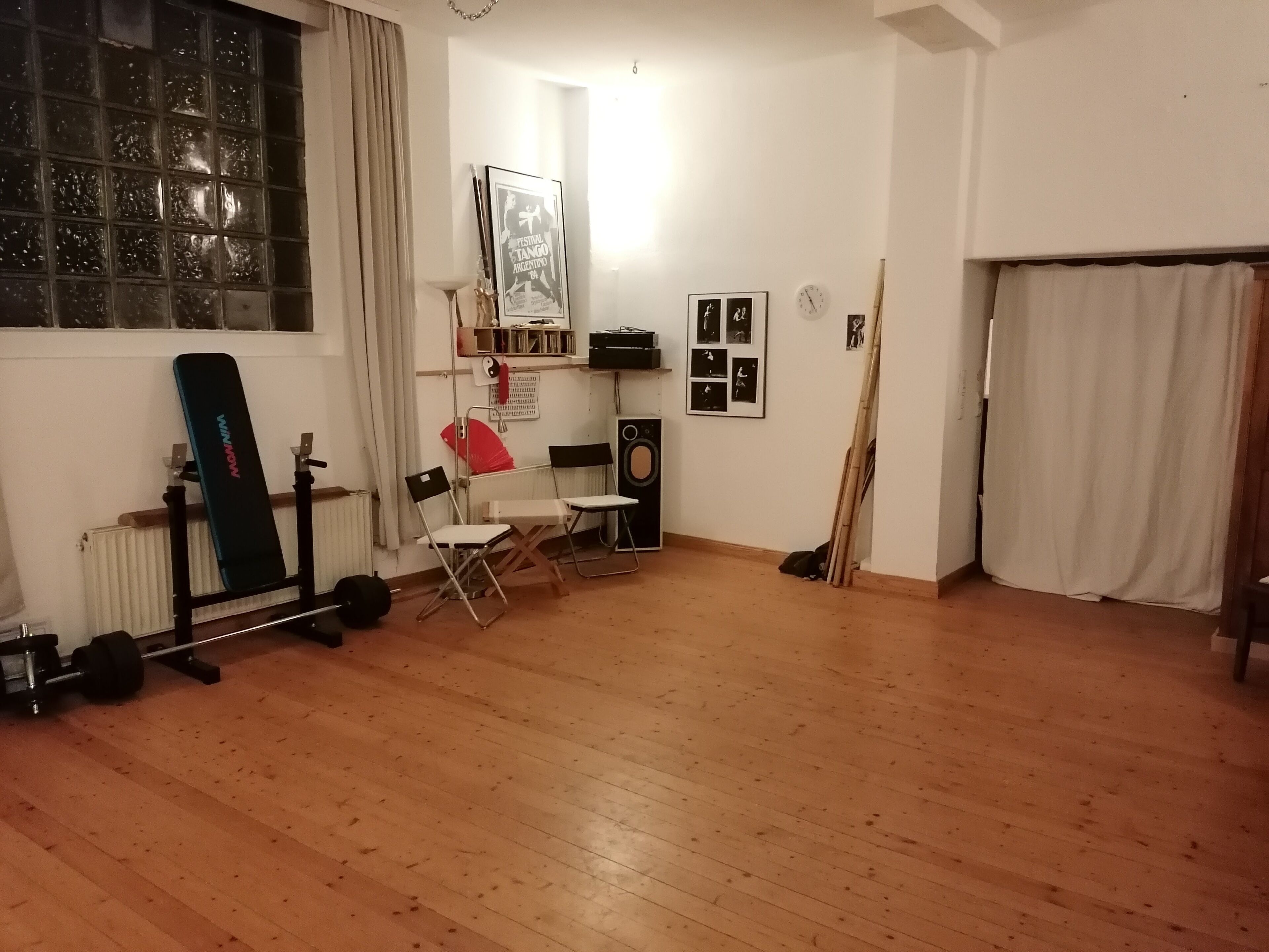 Een kamer met houten vloer, een raam met gordijnen, een radiator, een wandplank met diverse spullen, een lijst met een poster en een klok.