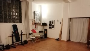 Een kamer met houten vloer, een raam met gordijnen, een radiator, een wandplank met diverse spullen, een lijst met een poster en een klok.