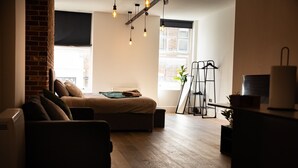 A cozy living room with a sofa, a coffee table, and a brick wall. There are hanging lights and a window with a view of the outside. - Brighton High Road Beach Studio (Brighton and Hove)