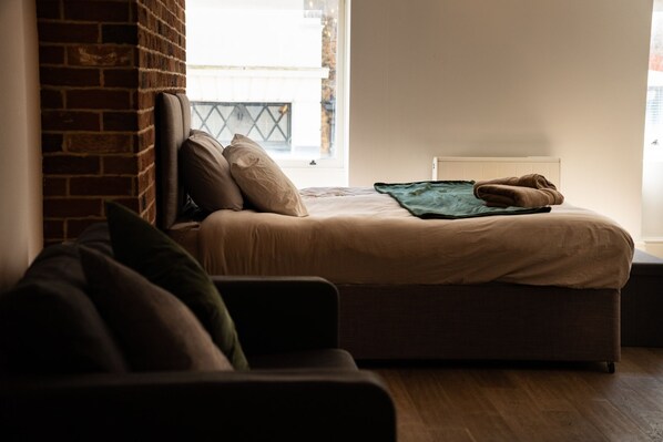 A bedroom with a bed, a brick wall, a window, and a view of the outside. - Brighton High Road Beach Studio (Brighton and Hove)