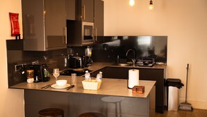 A modern kitchen with stainless steel appliances, a central island with stools, and a broom in the corner. - Brighton High Road Beach Studio (Brighton and Hove)