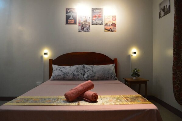 A neatly made bed with a pink bedspread and a rolled towel, a small bedside table with a plant, and wall-mounted light fixtures.