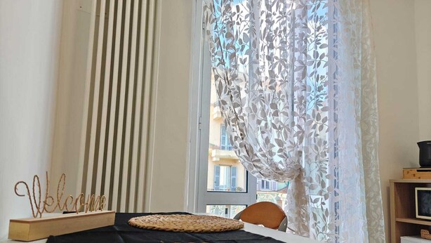 Habitación de hotel con cama, un cartel de 'Welcome', vista de edificios a través de cortinas traslúcidas y una pequeña mesa cubierta con un paño negro.