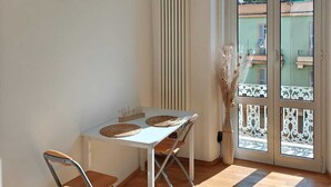 Una habitación con un reloj de pared de madera, una mesa con dos sillas, un radiador y vistas a edificios a través de un ventanal.