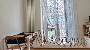Una habitación con una mesa de madera, una silla, un radiador, una ventana con cortinas florales y un cartel que dice 'Welcome'.