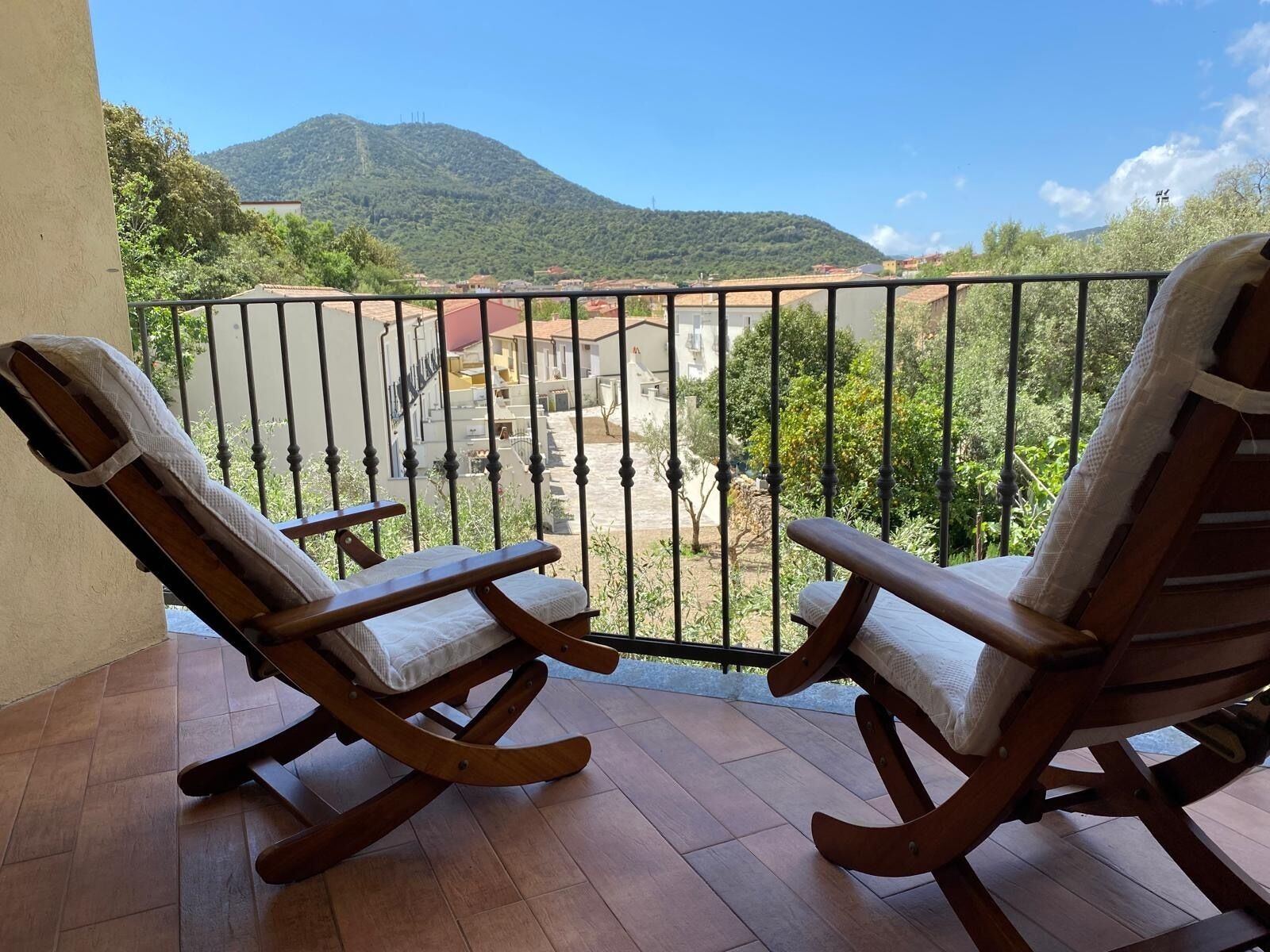 A balcony with two wooden rocking chairs and a metal railing, overlooking a hillside and buildings.