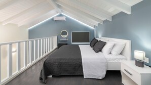 A bedroom with a sloped ceiling, a bed with a white headboard, a bedside table with a lamp, and a wall-mounted TV. - Golden sand 3 (Ialisos)