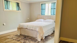 A bedroom with a bed, a window with curtains, a light switch, and a wooden floor. - Charming home on the lake! (Macy)
