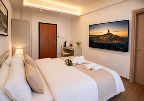 A hotel room with a bed, a desk, a chair, a TV, and a painting of a cityscape. - Central Domus (Matera)