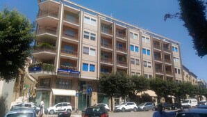 A multi-story building with balconies, a sign for 'Assicurazioni AXI INVESTIMENTI', and parked cars on the street. - Central Domus (Matera)